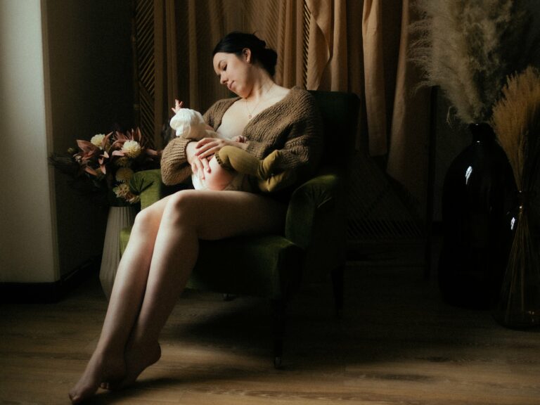 Maman allaitant son bébé assise dans un fauteuil vert en gilet tricot marron dans un décor studio cosy avec pampa et fleurs