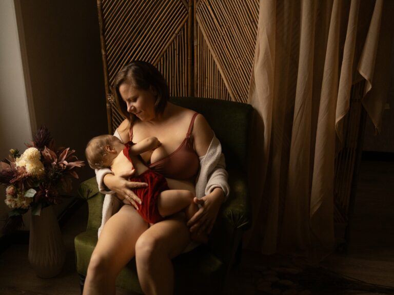 Maman allaitant son bébé en robe rouge assise dans un fauteuil vert en studio avec paravent en bambou et bouquet de fleurs séchées séance photo allaitement intimiste