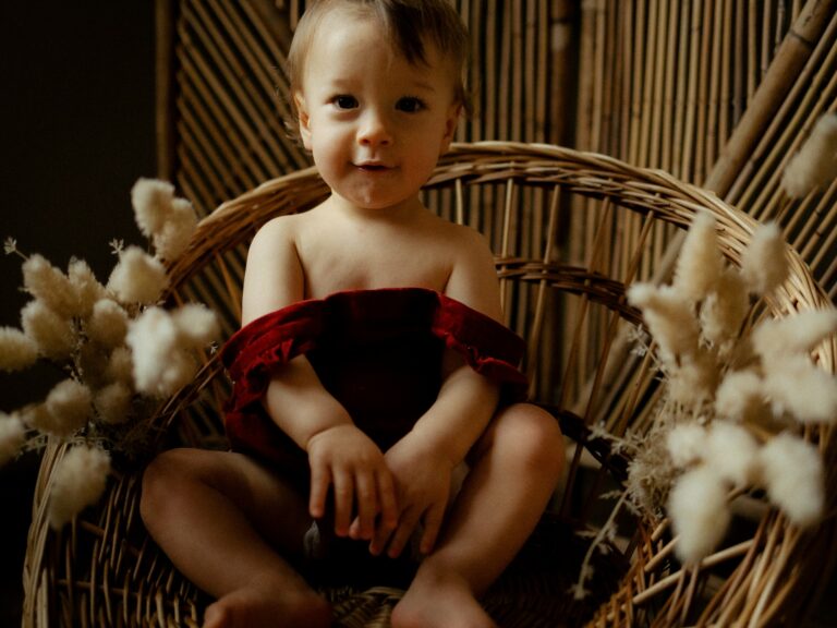 Bébé assis dans un fauteuil en rotin portant un bloomer rouge entouré de fleurs séchées bunny tails lors d'une séance photo portrait en studio
