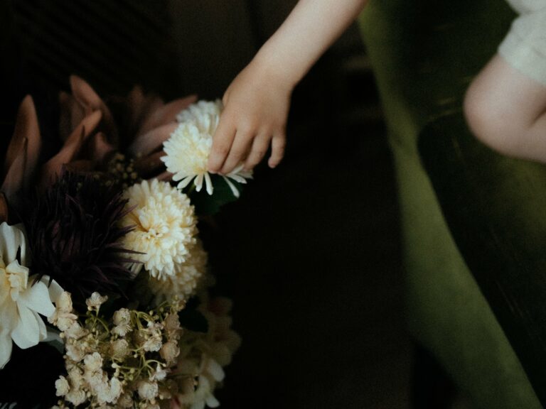 Petite main de bébé touchant délicatement un bouquet de fleurs avec dahlias blancs et violets lors d'une séance photo naissance artistique