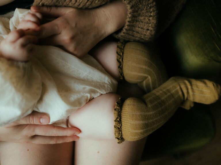 Détail pieds de bébé nouveau-né tenus par les mains de maman portant un pull en tricot marron séance photo naissance intimiste en lumière douce