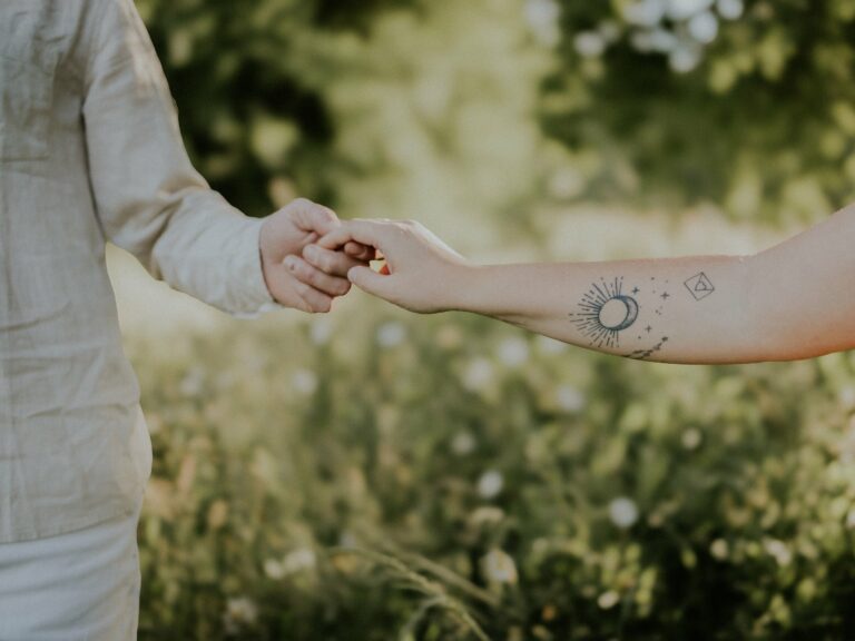 Détail des mains d'un parent en chemise lin et d'un enfant avec tatouage céleste sur l'avant-bras se touchant dans un décor de verdure lors d'une séance photo famille en extérieur
