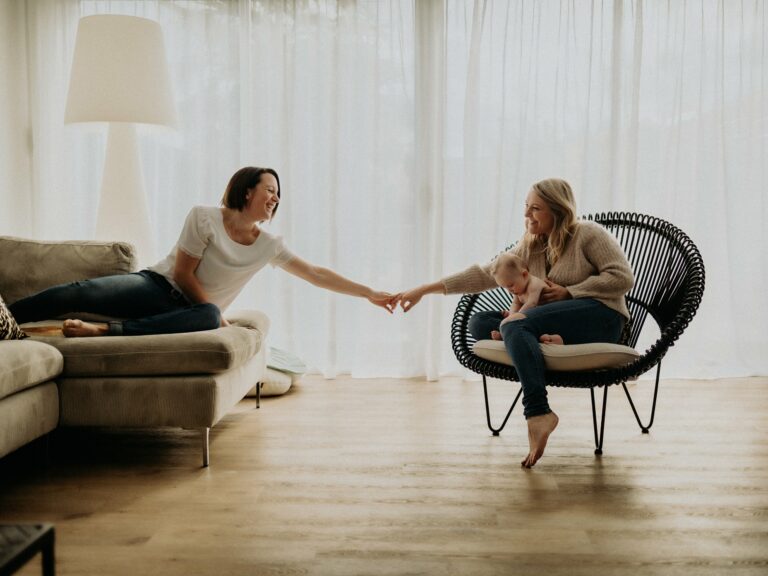 Deux mamans se tendant la main dans un salon lumineux l'une sur le canapé l'autre dans un fauteuil rotin avec bébé sur les genoux lors d'une séance photo famille lifestyle