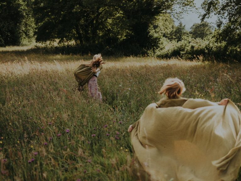 Deux enfants en robes courant librement dans une prairie d'herbes hautes et de fleurs sauvages avec arbres en arrière-plan lors d'une séance photo famille en extérieur
