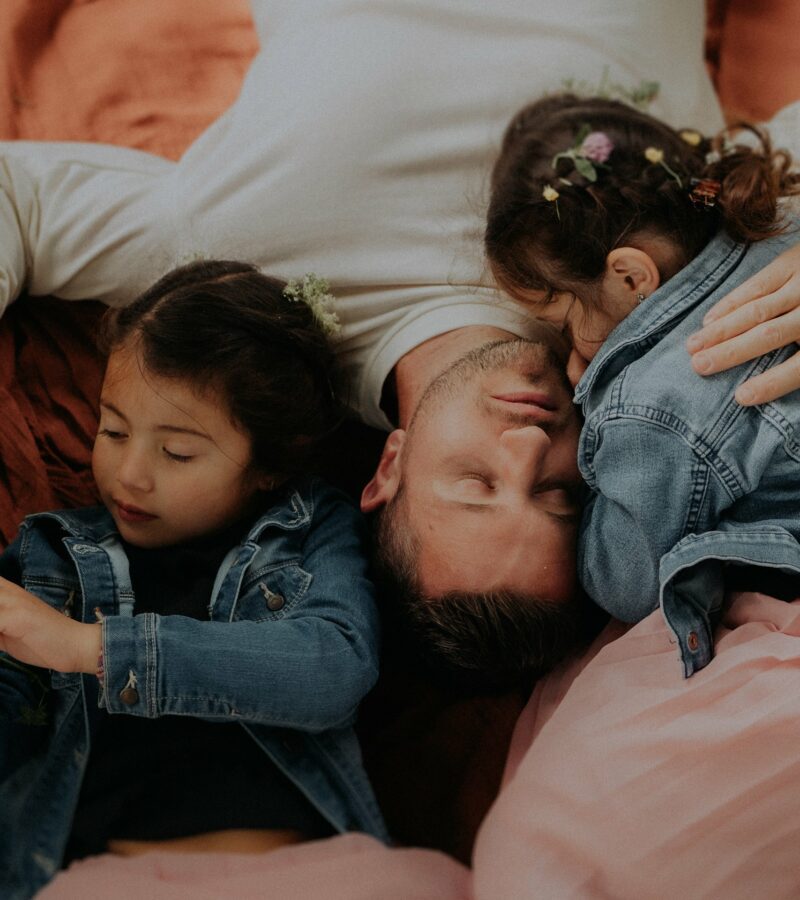 Papa allongé sur un plaid terracotta avec ses deux filles en veste jean et couronne de fleurs vue plongeante lors d'une séance photo famille en extérieur