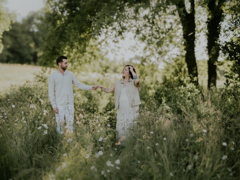 Couple attendant un bébé se tenant la main dans un champ fleuri en tenue blanche lors d'une séance photo grossesse en extérieur