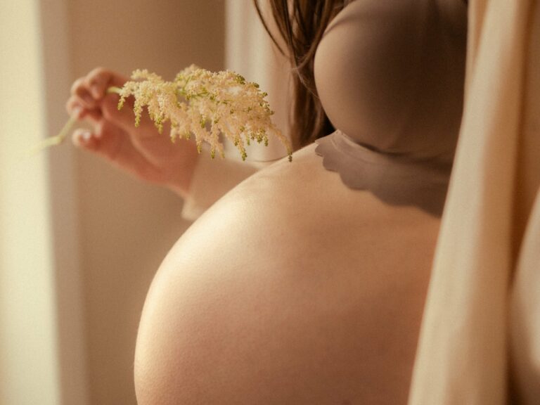 Gros plan sur un ventre rond de femme enceinte tenant une fleur blanche d'astilbe dans une lumière douce et chaude lors d'une séance photo grossesse en intérieur