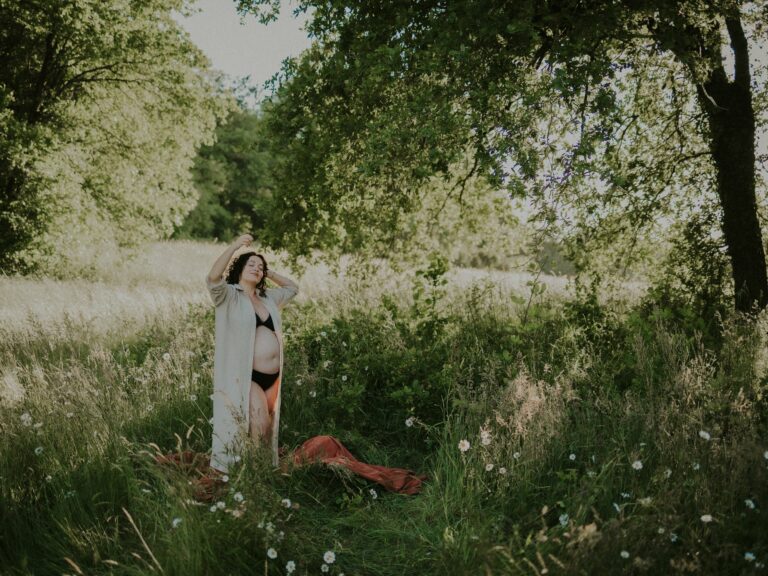 Femme enceinte debout dans un champ fleuri portant un long gilet ouvert avec ventre rond en pleine nature verdoyante lors d'une séance photo grossesse en extérieur