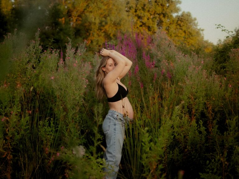 Femme enceinte blonde en brassière noire et jean posant dans un champ de fleurs roses et violettes lors d'une séance photo grossesse en extérieur