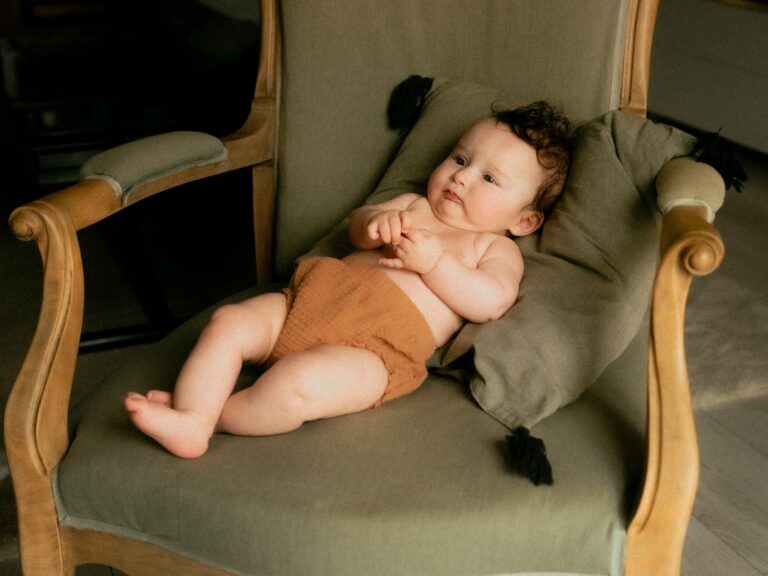 Bébé aux cheveux bouclés en bloomer terracotta allongé sur un fauteuil vintage vert olive lors d'une séance photo naissance à domicile