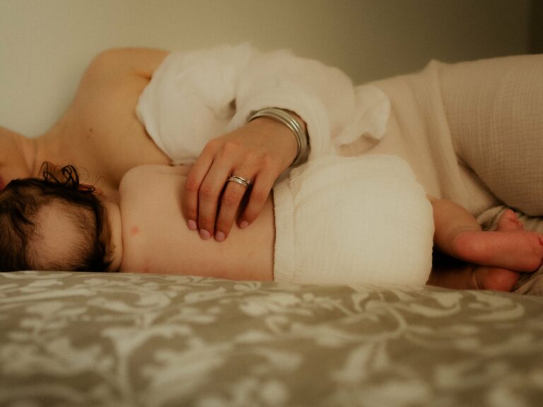 Maman allongée sur un lit avec son nouveau-né emmailloté dans un lange blanc main posée délicatement sur bébé avec bague et bracelets lors d'une séance photo naissance en intérieur