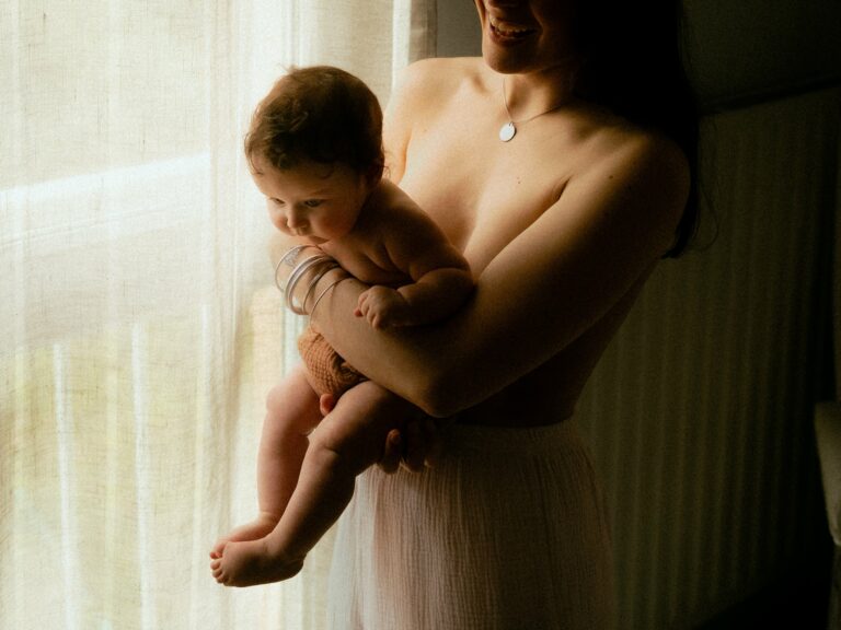 Maman portant son bébé dans les bras près d'une fenêtre en lumière douce lors d'une séance photo naissance intime