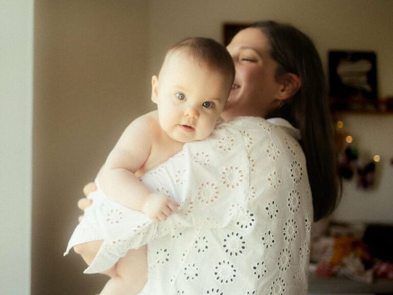 Maman portant son bébé sur l'épaule avec le bébé regardant la caméra en blouse blanche broderie anglaise lors d'une séance photo naissance à domicile