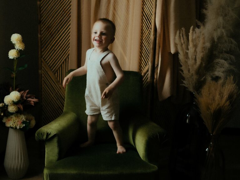 Petit garçon souriant debout sur un fauteuil vert en studio avec paravent en bois et herbe de pampa lors d'une séance photo portrait enfant
