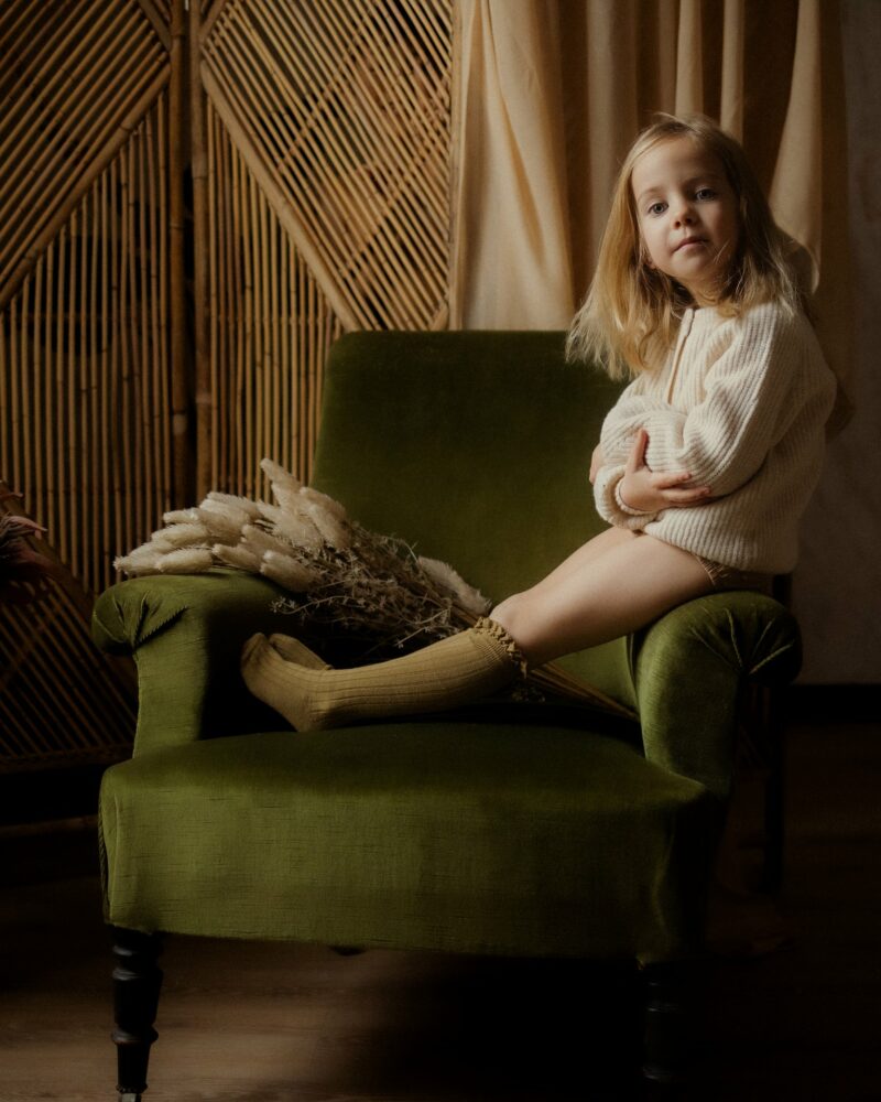 Petite fille blonde assise sur un fauteuil vert en studio portant un pull blanc avec décor bambou pampa et fleurs lors d'une séance portrait enfant