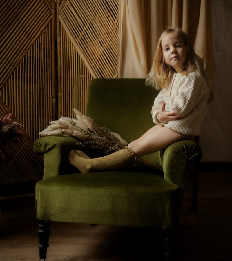 Petite fille blonde assise sur un fauteuil vert en studio portant un pull blanc avec décor bambou pampa et fleurs lors d'une séance portrait enfant