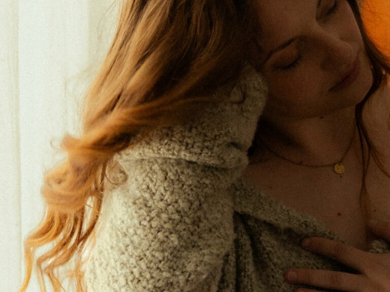 Portrait intimiste d'une femme aux cheveux roux les yeux fermés portant un gilet bouclé crème et un collier doré en lumière chaude en studio