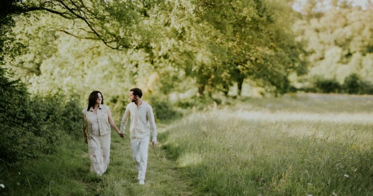 Couple se promenant main dans la main sur un chemin de campagne verdoyant habillés en blanc et crème lors d'une séance photo en extérieur en lumière dorée