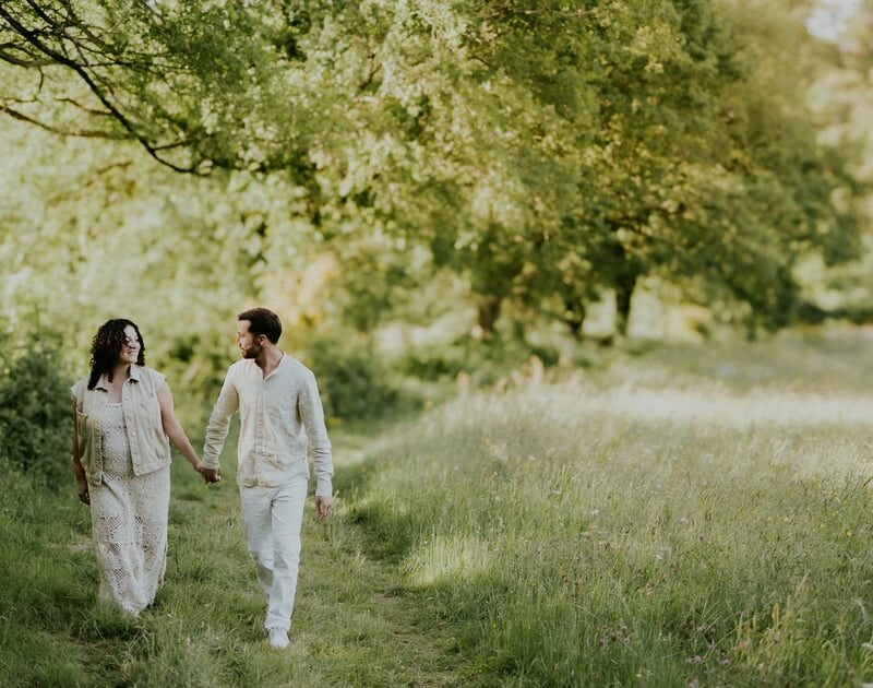 Couple se promenant main dans la main sur un chemin de campagne verdoyant habillés en blanc et crème lors d'une séance photo en extérieur en lumière dorée