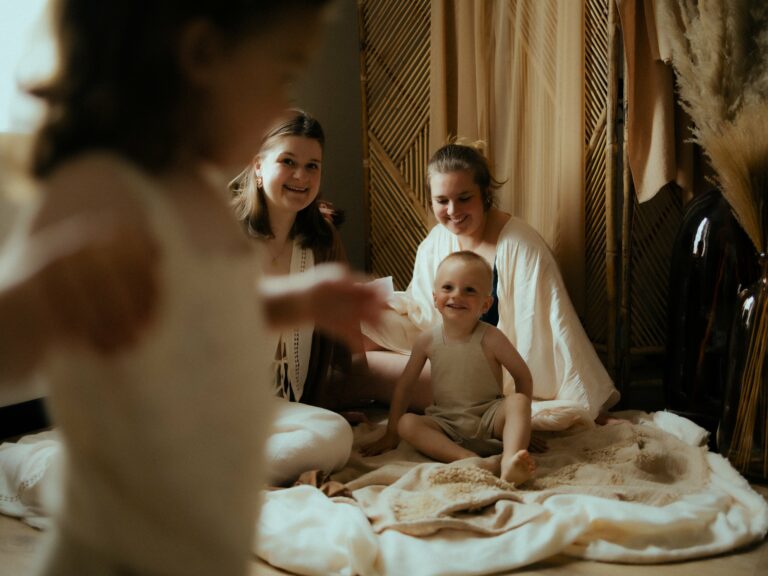 Deux mamans souriantes assises au sol avec leur bébé souriant et enfant au premier plan lors d'une séance photo famille en studio avec paravent bambou et pampa