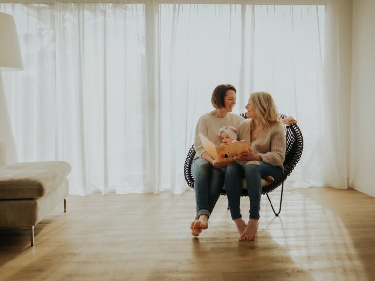 Deux mamans souriantes assises dans un fauteuil en rotin avec leur bébé lisant un livre ensemble lors d'une séance photo famille en studio lumineux avec rideaux blancs