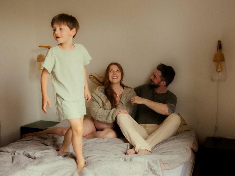 Enfant sautant joyeusement sur le lit avec ses parents assis qui rient lors d'une séance photo famille lifestyle à domicile spontanée et chaleureuse
