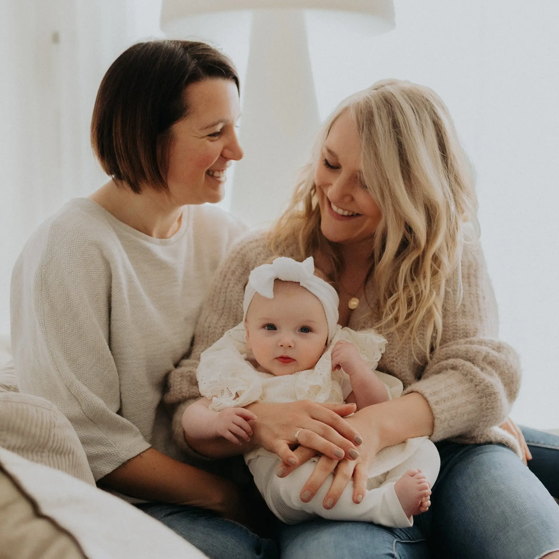 Deux mamans souriantes avec leur bébé sur le canapé lors d'une séance famille homoparentale à domicile - Melissa Jai Photographe
