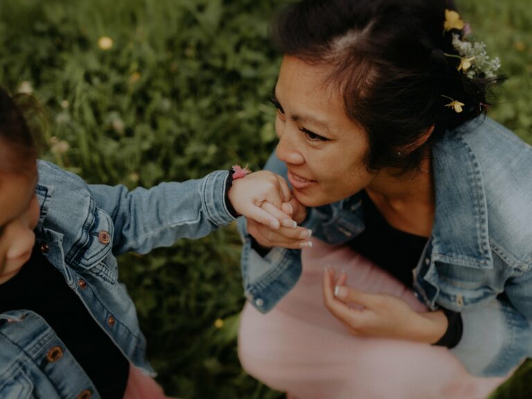 Maman embrassant tendrement la main de son enfant lors d'une séance photo famille en extérieur dans l'herbe avec fleurs dans les cheveux et vestes en jean assorties