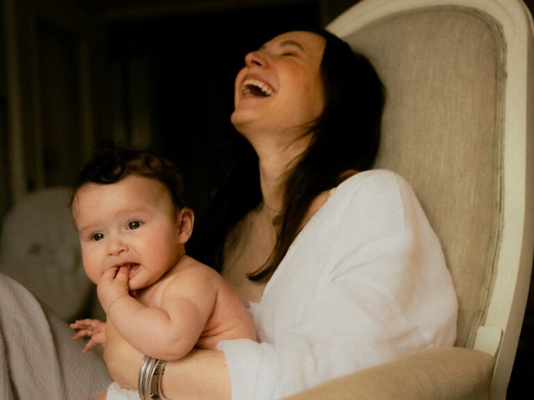 Maman éclatant de rire avec son bébé sur les genoux dans un fauteuil lors d'une séance photo famille lifestyle intime en lumière douce et sombre