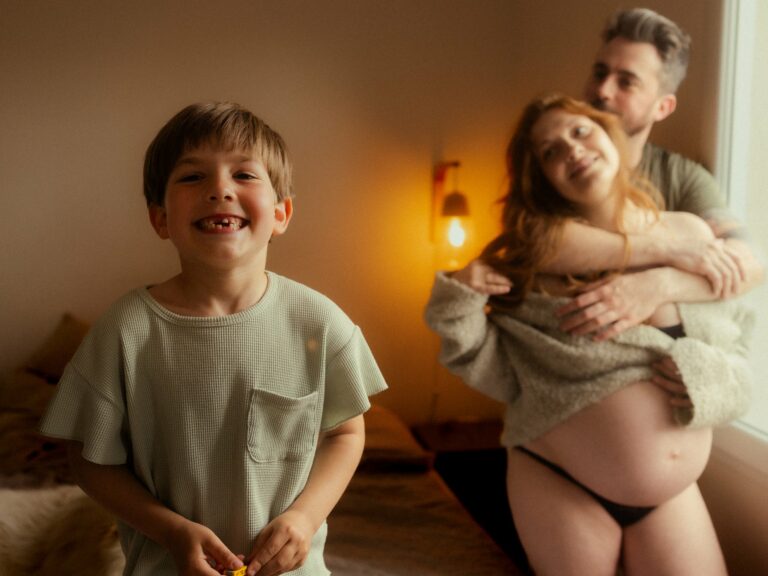 Enfant souriant au premier plan avec ses parents en arrière-plan papa enlacant maman enceinte lors d'une séance photo grossesse en famille en studio avec lumière chaude