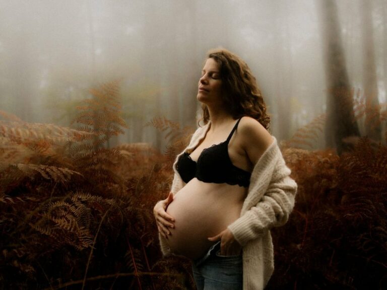 Femme enceinte les yeux fermés tenant son ventre dans une forêt brumeuse entourée de fougères automnales portant un gilet en laine et lingerie noire séance photo grossesse extérieur