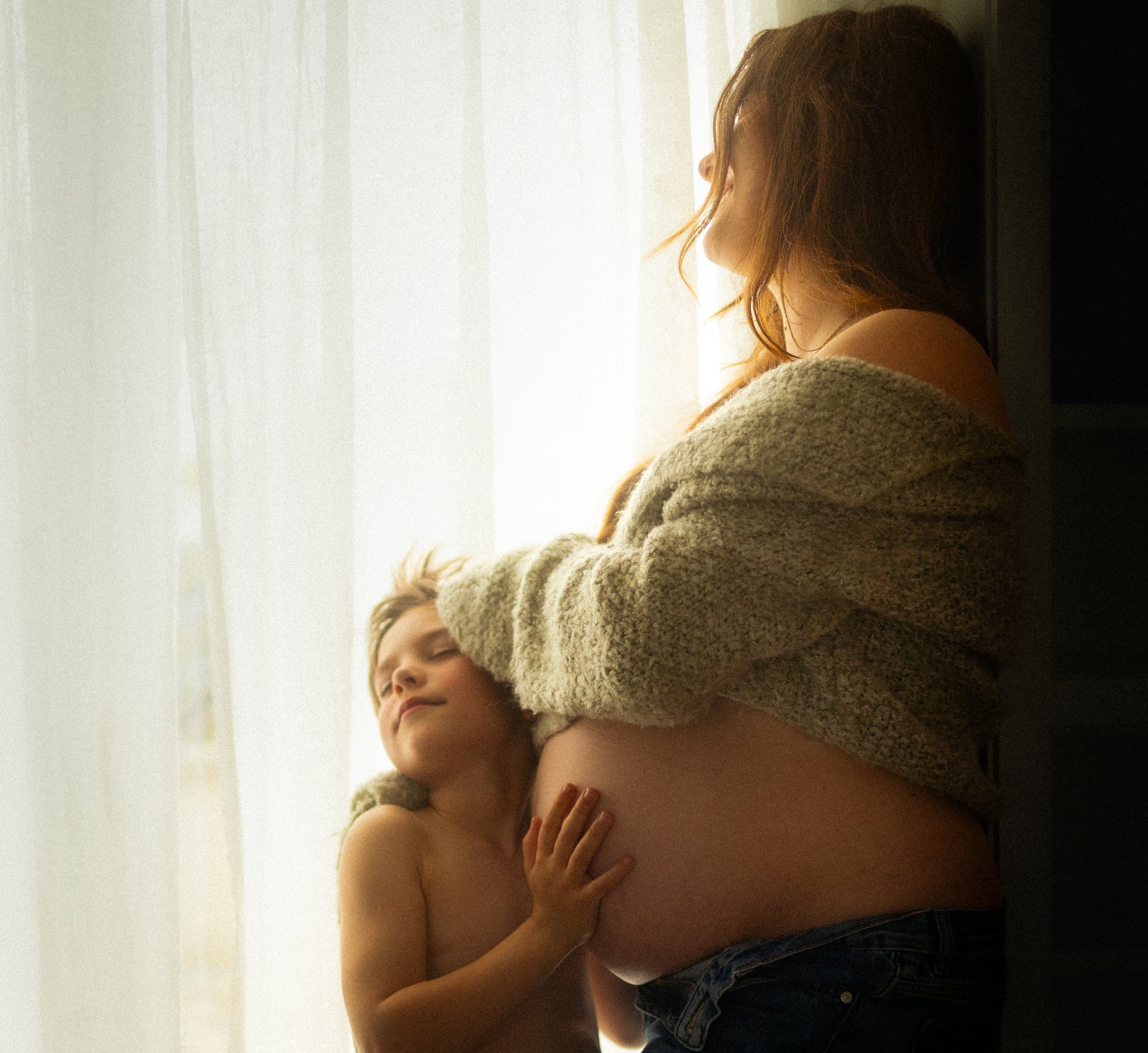 Femme enceinte de profil regardant par la fenêtre en lumière dorée lors d'une séance grossesse - Melissa Jai Photographe