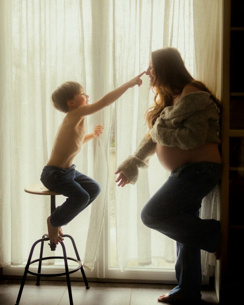Maman et enfant en contre-jour devant un rideau lors d'une séance grossesse à domicile - Melissa Jai Photographe