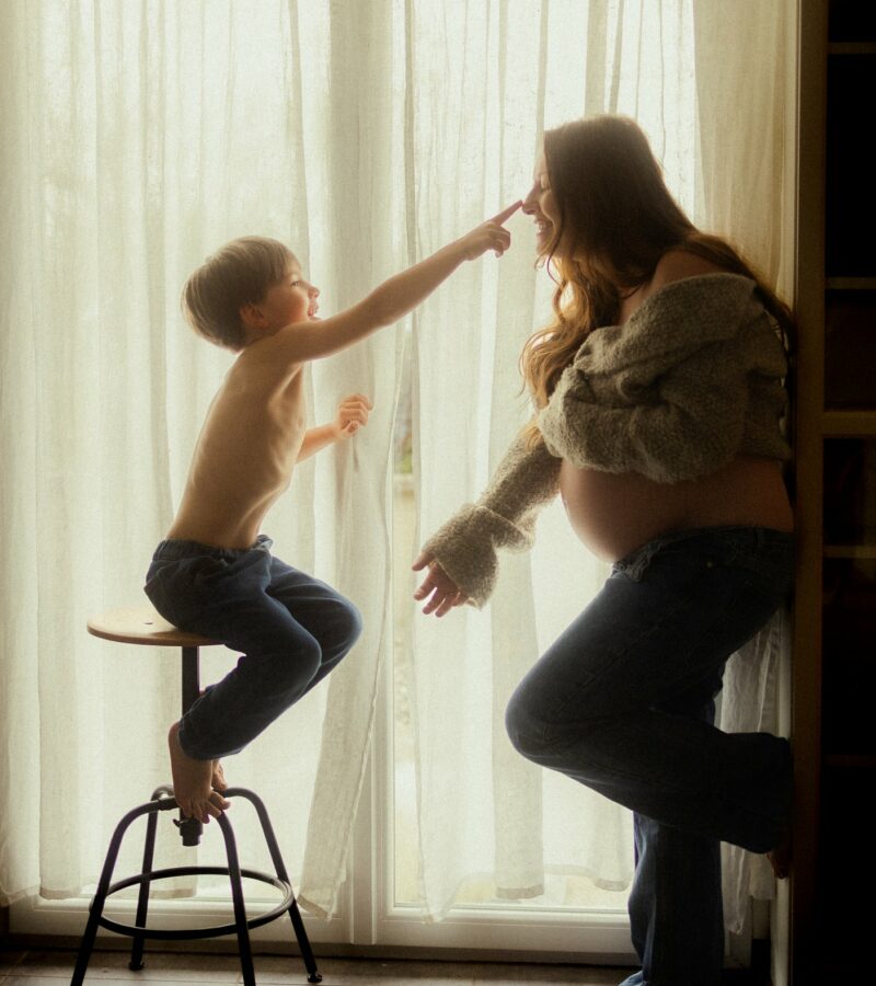 Maman et enfant en contre-jour devant un rideau lors d'une séance grossesse à domicile - Melissa Jai Photographe