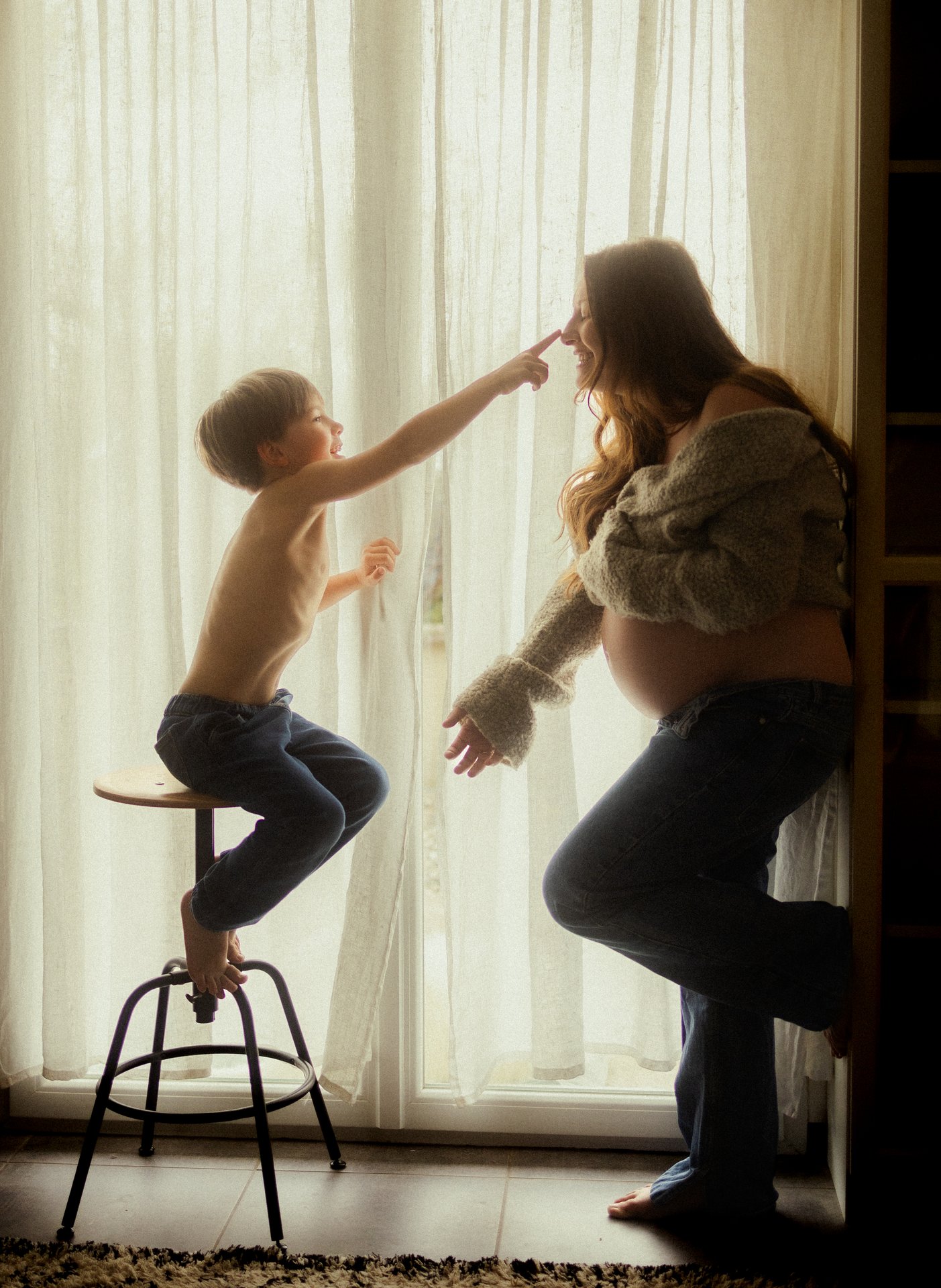 Maman et enfant en contre-jour devant un rideau lors d'une séance grossesse à domicile - Melissa Jai Photographe