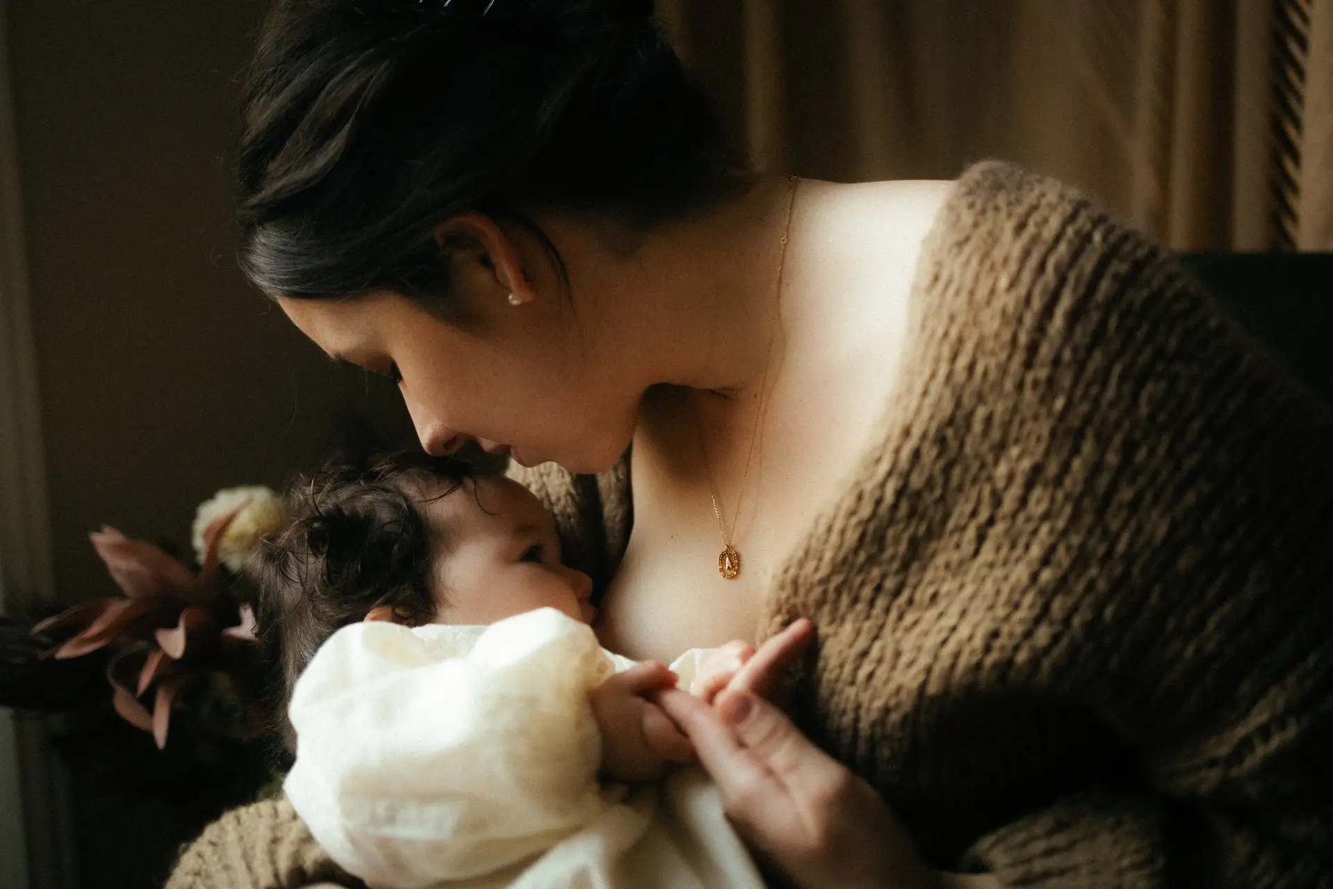 Maman tenant son nouveau-né contre elle avec un collier doré lors d'une séance naissance en lumière douce - Melissa Jai Photographe