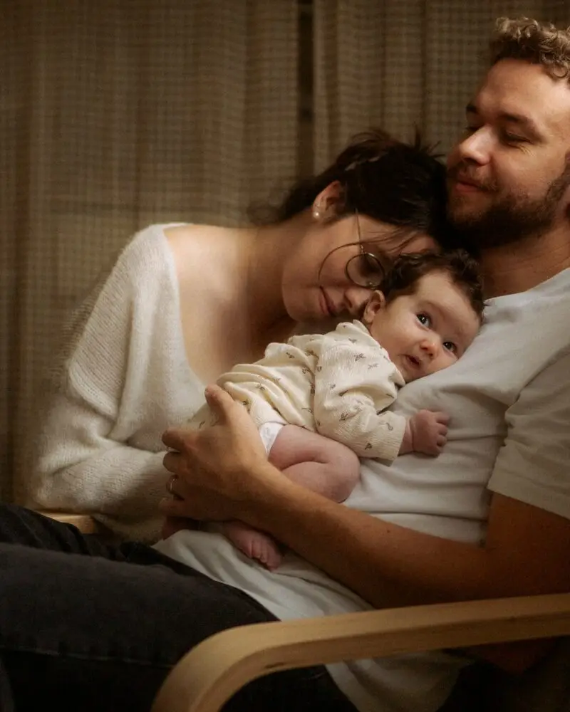 Papa tenant son nouveau-né dans un fauteuil à bascule avec maman blottie contre eux lors d'une séance naissance à domicile - Melissa Jai Photographe