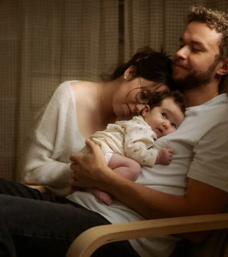 Papa tenant son nouveau-né dans un fauteuil à bascule avec maman blottie contre eux lors d'une séance naissance à domicile - Melissa Jai Photographe