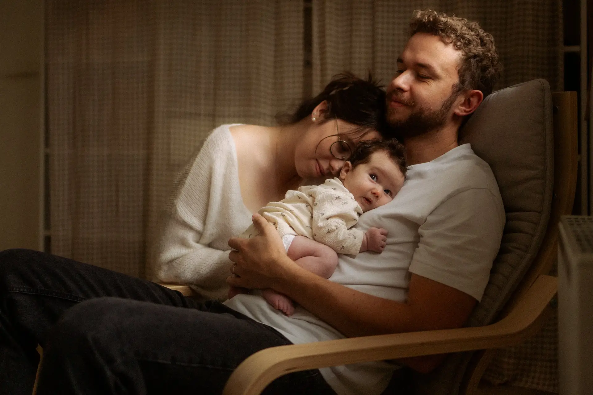Papa tenant son nouveau-né dans un fauteuil à bascule avec maman blottie contre eux lors d'une séance naissance à domicile - Melissa Jai Photographe