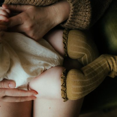 Détail pieds de bébé nouveau-né tenus par les mains de maman portant un pull en tricot marron séance photo naissance intimiste en lumière douce
