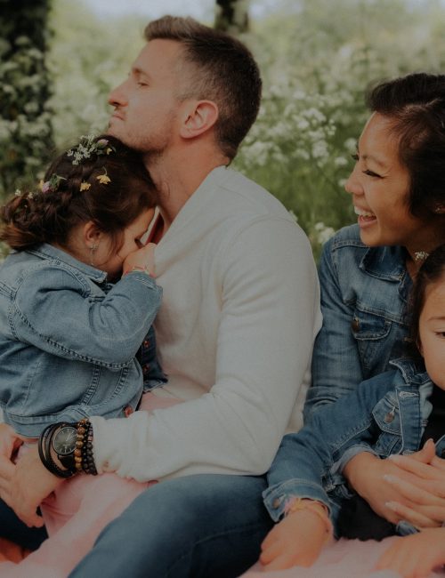 Famille avec papa maman et deux petites filles en vestes jean avec fleurs dans les cheveux assise dans un champ fleuri lors d'une séance photo famille en extérieur nature