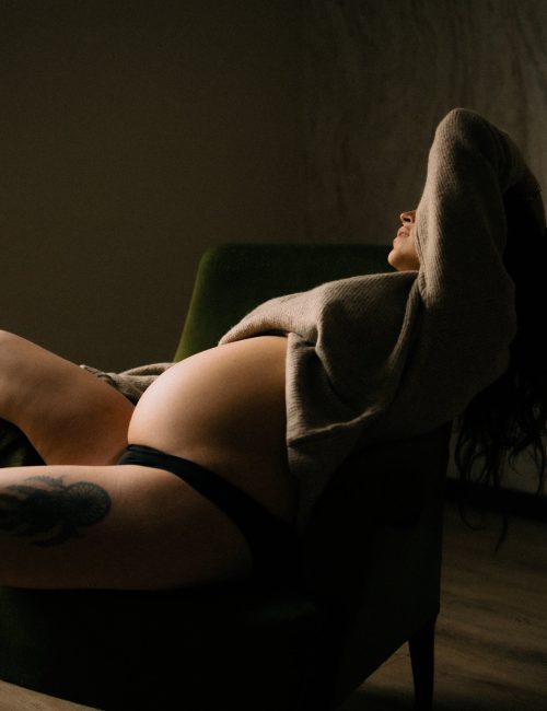Femme enceinte allongée sur un fauteuil vert en gilet tricot avec ventre rond en clair-obscur lors d'une séance photo grossesse boudoir en studio