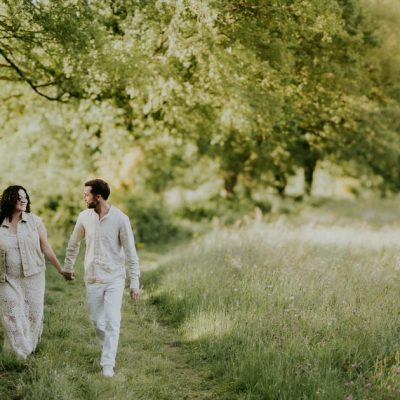 Couple se promenant main dans la main sur un chemin de campagne verdoyant habillés en blanc et crème lors d'une séance photo en extérieur en lumière dorée