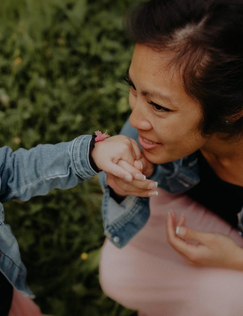 Maman embrassant tendrement la main de son enfant lors d'une séance photo famille en extérieur dans l'herbe avec fleurs dans les cheveux et vestes en jean assorties