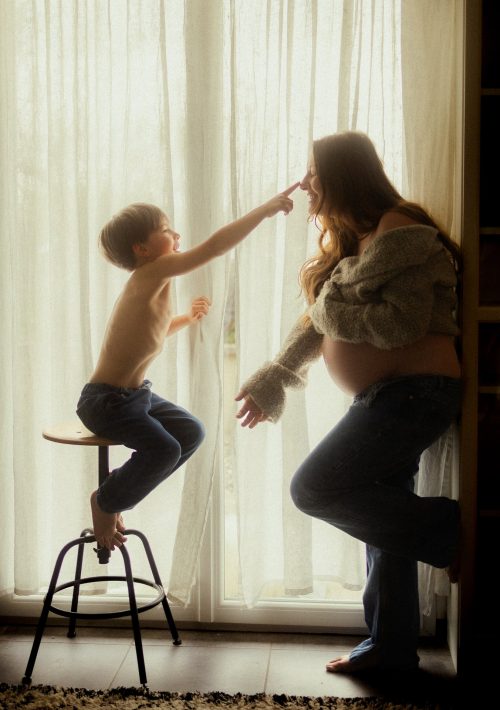 Maman et enfant en contre-jour devant un rideau lors d'une séance grossesse à domicile - Melissa Jai Photographe