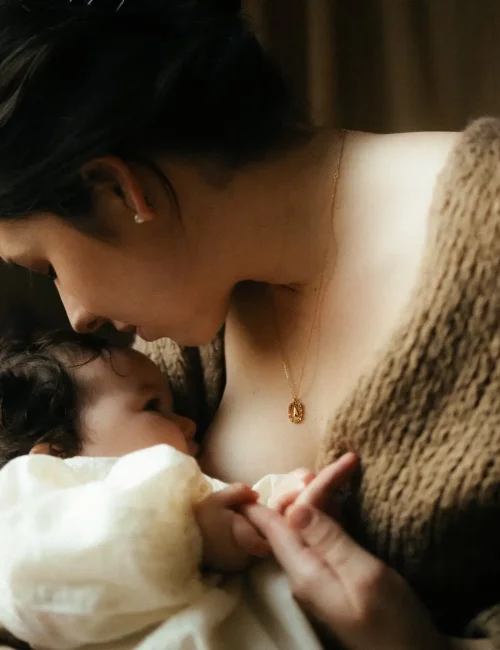 Maman tenant son nouveau-né contre elle avec un collier doré lors d'une séance naissance en lumière douce - Melissa Jai Photographe